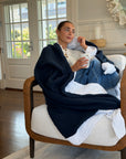 A woman sits in a white armchair wrapped in a navy blue and snow Gray Heron muslin blanket, holding a white mug in a bright living room.