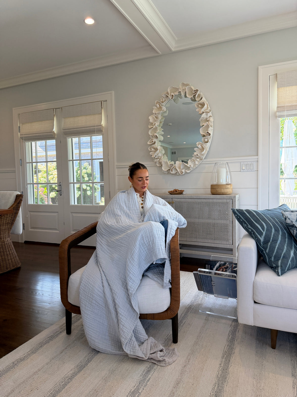 A woman sits in a wooden armchair wrapped in a Mist All-season Gray Heron t inside a bright, modern living room with white walls, a shell-mirrored wall mirror, and a white sofa with blue pillows.