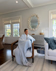 A woman sits in a wooden armchair wrapped in a Mist All-season Gray Heron t inside a bright, modern living room with white walls, a shell-mirrored wall mirror, and a white sofa with blue pillows.