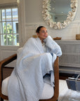 A woman sits in a wicker armchair wrapped in a mistmuslin blanket, in a bright, modern living room with a large decorative mirror on the wall.